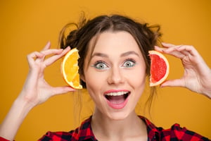 Close up of excited happy young woman holding slices of orange and grapefruit over yellow background